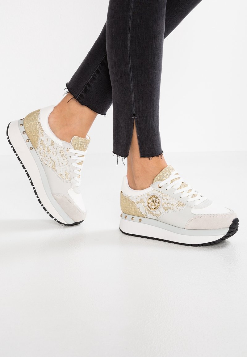 Hvide sneakers med guld accenter, snøre detaljer og en chunky sål. Har et logo på siden og dekorative nitter langs designet.