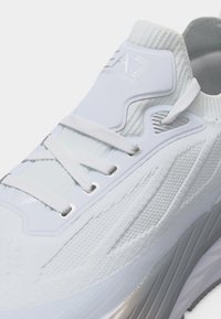 Λευκό πλεκτό αθλητικό παπούτσι με ενσωματωμένα κορδόνια, πλαστικές τρύπες και λογότυπο στη γλώσσα και στην πλαϊνή πλευρά.