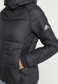 Sort polstret jakke med hætte, prydet med skrå sømme, lynlåslukning og elastiske manchetter. Logo på venstre ærme.