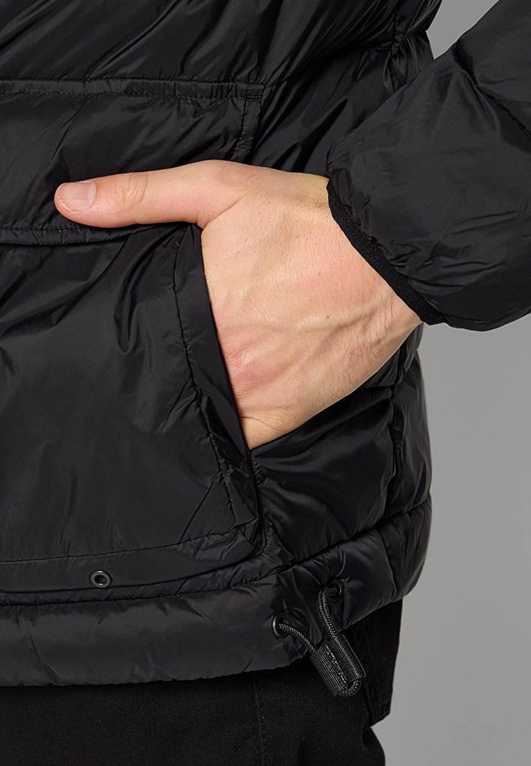 Hand gedeeltelijk in de zak van een zwarte gewatteerde jas, toont de textuur van de jas en de verstelbare koord aan de zoom.