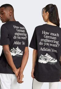 Svart oversize t-shirt med stor vit text på ryggen och en grafik av en sneaker. Korta ärmar och avslappnad passform.