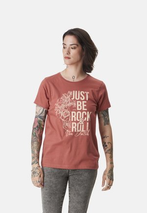 Femme tatouée portant un T-shirt couleur rouille avec un dessin de crâne et de fleurs ainsi que le texte "JUST BE ROCK & ROLL Von Dutch", associé à un jean gris.