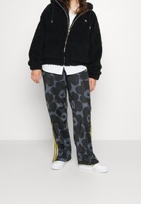 Sudadera de polar negra con cremallera dorada. Pantalones grises con estampado floral que presentan rayas laterales amarillas. Zapatillas blancas. Camiseta blanca debajo.