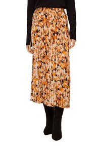 Kvinna som bär en plisserad midikjol med ett mönster i orange, svart och vitt, svart långärmad topp och svarta stövlar.