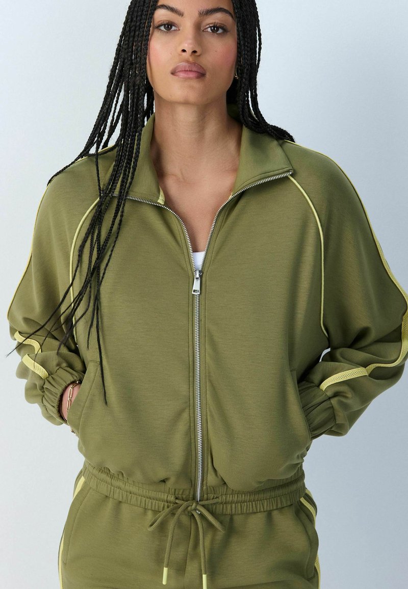 Jeune femme avec de longues tresses portant une veste zippée vert olive et un pantalon assorti, mains dans les poches, fond neutre.