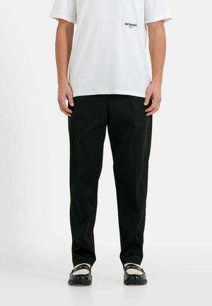 Schwarze Hose mit geradem Schnitt und glatter Textur, kombiniert mit einem locker sitzenden weißen T-Shirt, das ein kleines Logo auf der Brust trägt.