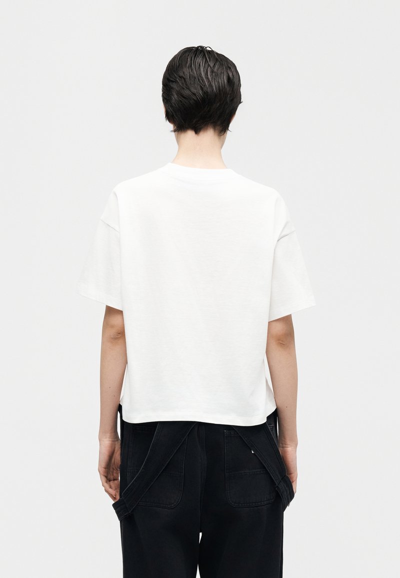 Person mit kurzen dunklen Haaren, von hinten gesehen, trägt ein lockeres weißes Kurzarmhemd und schwarze Hosen mit herunterhängenden Hosenträgern.