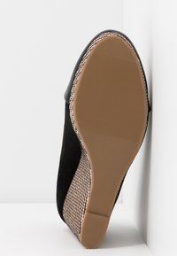 Chaussure en suède noir avec un côté texturé et tissé et une semelle lisse marron. Le talon est rond et s'intègre parfaitement au corps de la chaussure.