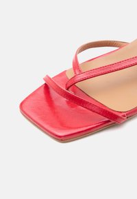 Κόκκινο σανδάλι ανοιχτής μύτης με τετράγωνο μπροστινό μέρος και δύο λεπτά λουράκια που διασταυρώνονται πάνω από το πόδι, δείχνοντας απαλό δέρμα και μπεζ εσωτερική σόλα.