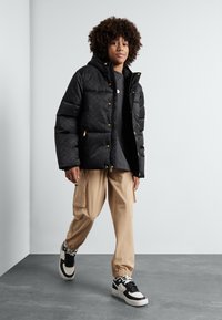 Sort pufferjakke med guldfarvet hardware, hætte og quiltet mønster. Båret med beige cargo-bukser og sorte sneakers med hvide detaljer.