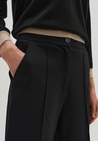 Black tailored pants with pleats, side pockets, and a button closure, made from a smooth fabric. A ribbed beige cuff is partially visible.