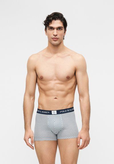 Tânăr cu păr închis la culoare, purtând boxeri Polo Ralph Lauren cu model alb, stând în picioare în fața unui fundal simplu, deschis la culoare.