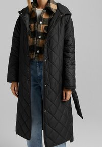 Czarny pikowany długi płaszcz z kapturem, z zatrzaskami i paskiem. Pod spodem widoczna jest koszula w kratę w odcieniach brązu i czerni.