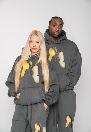 Two models wearing matching dark gray hoodies and sweatpants with yellow-black abstract mushroom designs, posing against a plain background.