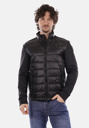 Hombre con cabello rizado que lleva una chaqueta negra acolchada con bolsillos con cremallera y pantalones vaqueros azules, de pie con una mano en el bolsillo.