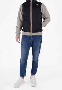 Mann in einer schwarzen Steppweste über einem beigen Sweatshirt, blauen Jeans und weißen Sneakers, steht mit einer Hand in der Tasche.