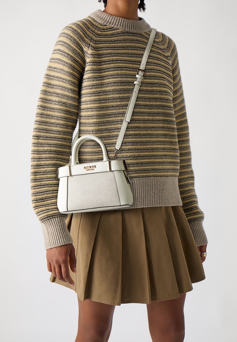 Personne portant un pull en maille rayé beige et jaune et une jupe plissée beige, tenant un sac à main blanc Guess avec une poignée supérieure et une bandoulière.