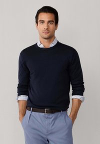 Mężczyzna o ciemnych włosach, ubrany w granatowy sweter na paskowaną koszulę i jasnoniebieskie spodnie, stoi z rękami w kieszeniach na jednolitym tle.