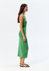 Grünes, ärmelloses, knielanges Kleid mit tailliertem Oberteil und glatter Textur. Kombiniert mit flachen Sandalen mit Riemendesign.