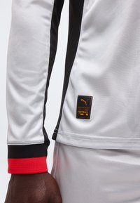 Αθλητική μπλούζα μακρυμάνικη με μαύρες και κόκκινες μανσέτες, μαύρη πλαϊνή ρίγα και μικρό patch με το λογότυπο Puma «Authentic Licensed Football» κοντά στη μέση.