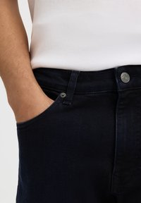 Persona que lleva vaqueros azul oscuro y una camisa blanca con la mano izquierda colocada dentro del bolsillo delantero de los vaqueros.