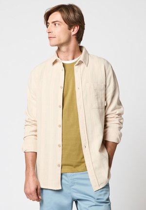 Homme portant une chemise crème boutonnée par-dessus un t-shirt moutarde et un pantalon bleu clair, regardant vers sa gauche avec une main dans la poche.