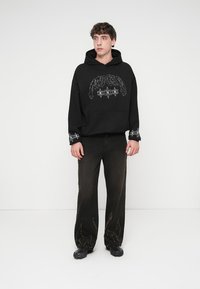 Μαύρο βαμβακερό φούτερ με κουκούλα, χαλαρή εφαρμογή και τσέπη μπροστά με γραφικό τύπωμα. Συνδυασμένο με σκούρο, άνετο παντελόνι με κεντημένες λεπτομέρειες.