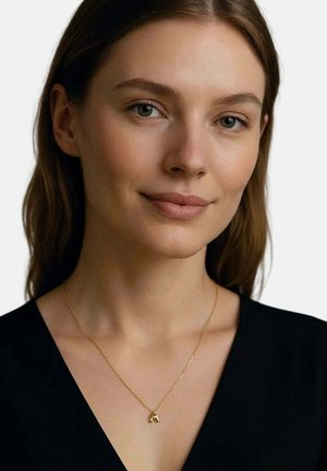 Giovane donna con capelli castani che indossa una collana con pendente dorato e una maglia nera, sorridendo dolcemente su uno sfondo chiaro e uniforme.