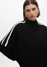 Černý svetr s vysokým límcem, bílými postranními pruhy na rukávech a volným střihem. Měkká textura se zrolovanými rukávy. Diskrétní logo.