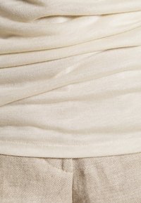 Béžový, lehký, žebrovaný top s měkkou texturou a vícevrstvým designem. Spárován se světle béžovými kalhotami z texturované tkaniny s jemným vzorem.