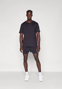 Čierne bavlnené tričko s krátkymi rukávmi v kombinácii s čiernymi športovými šortkami. Model má na sebe biele tenisky a ponožky, stojí pred jednofarebným pozadím.