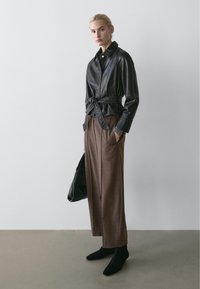 Kvinde iført en sort læderjakke med bælte, brune vidde bukser, sorte sko, der holder en sort taske, stående op ad en ensfarvet væg.