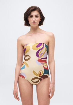 Kobieta z krótkimi brązowymi włosami, ubrana w jednoczęściowy strój kąpielowy bez ramiączek z kolorowym abstrakcyjnym wzorem liści na beżowym tle.