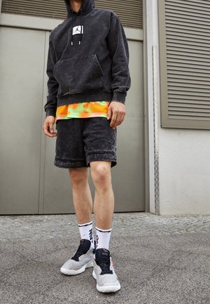 Person iført en sort hoodie med Jordan-logo, neongul orange skjorte under, sorte shorts, hvide sokker med sort tekst og grå sneakers står udendørs.