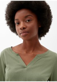 Jonge vrouw met een natuurlijke afro-haarstijl, gekleed in een lichtgroene V-hals top, kijkt rustig in de camera tegen een witte achtergrond.