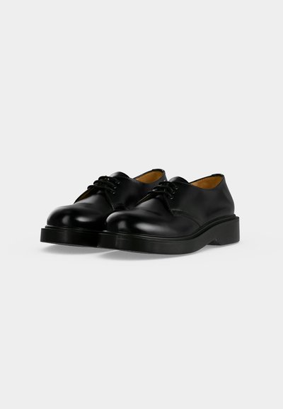 Paire de chaussures habillées en cuir noir à lacets avec semelles épaisses sur fond gris clair uni.