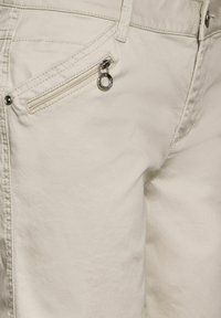 Light beige pants featuring a zippered pocket on the front, smooth fabric texture, and metal hardware accents for closure.