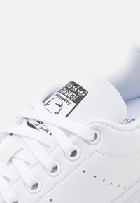 Sneaker bianca in pelle con lacci bianchi piatti, dettagli perforati sui lati e un'etichetta di marca nera sulla linguetta.