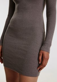 Närbild av en person som bär en figurnära, ribbad, långärmad klänning i taupe med kort nederdel, som visar delar av överkroppen, armarna och händerna.