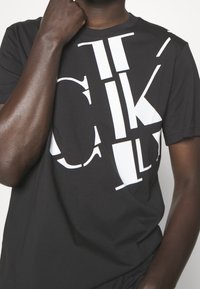 Svart bomulls t-shirt med stor vit typografi på framsidan. Korta ärmar och rundad halsringning. Slät textur.