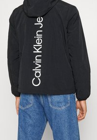 Chaqueta con capucha negra que presenta una textura suave y el logotipo en texto blanco "Calvin Klein Jeans" en vertical en la parte posterior. Combinada con jeans de mezclilla azules.