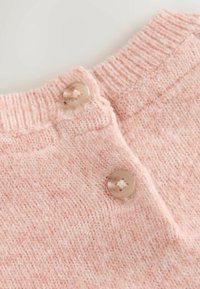 Helles rosa gestricktes Material mit einer strukturierten Oberfläche. Zwei runde, neutralfarbene Knöpfe sind am Ausschnitt sichtbar.