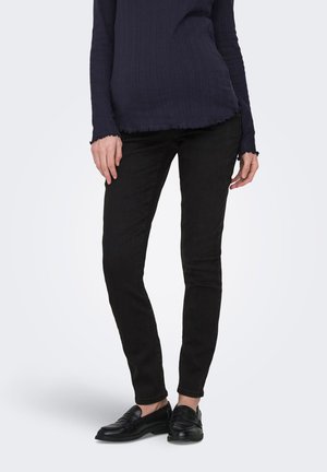 Person iført sorte slim fit bukser, sorte loafers og en mørkeblå ribbet langærmet trøje, der står mod en lys baggrund.