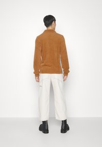 Brun, fluffig tröja med hög krage, bärs tillsammans med vita vida byxor och svarta ankelstövlar, sedd bakifrån.