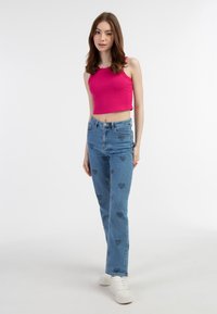 Top corto rosa e jeans blu a vita alta con motivi a cuore. Modello con scarpe bianche, in piedi su uno sfondo chiaro.