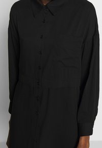 Camicia nera con bottoni, colletto, maniche lunghe, una tasca sul petto e dettaglio in vita arricciato. Tessuto dalla finitura liscia.