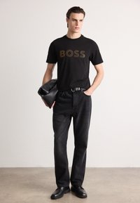 Juodas marškinėliai su auksiniu "BOSS" logotipu, derinami su tamsiai džinsais ir juodais batais. Modeliai ant paprasto fono, laikantys juodą rankinę.
