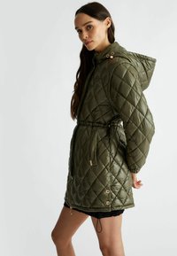 Giacca puffer trapuntata verde oliva con cappuccio regolabile, vita aderente e bottoni a scatto in tonalità oro, caratterizzata da una texture di tessuto liscia e lucida.