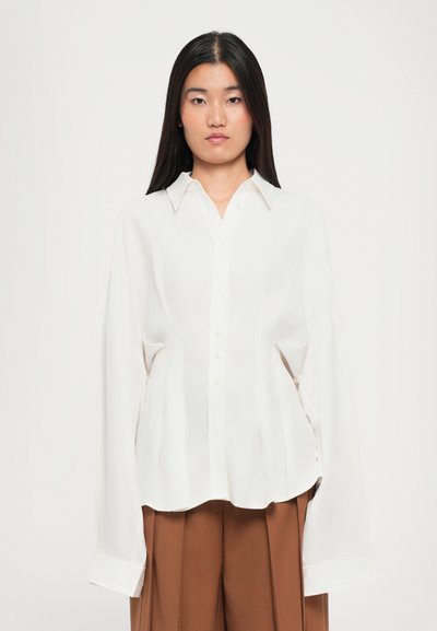 Blouse légère de couleur crème avec un col classique, des boutons à l'avant et des manches longues ; présente une coupe décontractée et de légères plis.