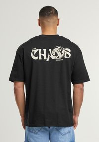 Black cotton t-shirt with a large white “CHAOS” graphic on the back and the text “FREE YOUR MIND,” short sleeves, and a relaxed fit.